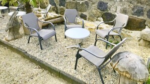 Un espace extérieur aménagé avec des chaises en osier et une table ronde, entouré d’un mur de pierre et d’un sol en gravier.