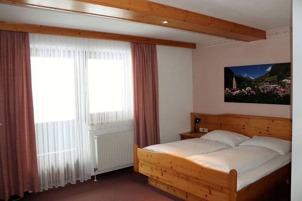 A bedroom with a wooden bed, a radiator, a window with sheer curtains, and a painting on the wall.