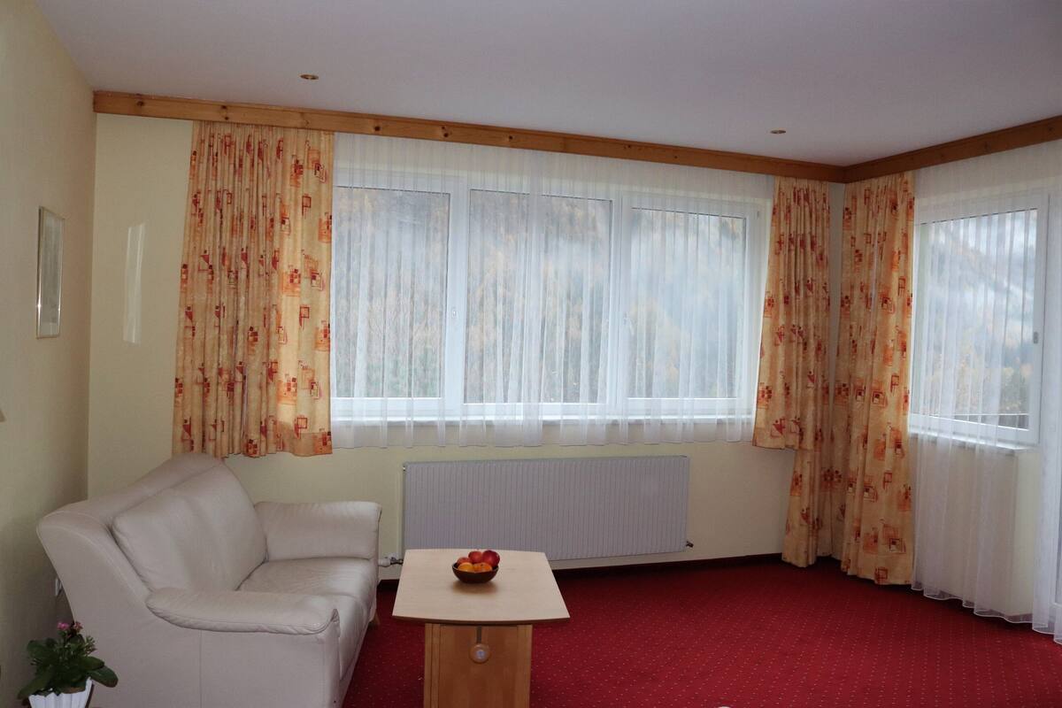 A room with a red carpet, a white sofa, a small wooden table with a bowl of fruit, and a radiator.