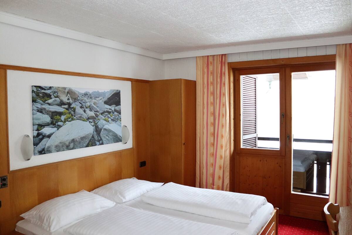 A hotel room with a wooden headboard, a bed with white linens, a painting of rocks, a wooden door with a window, and a red carpet.