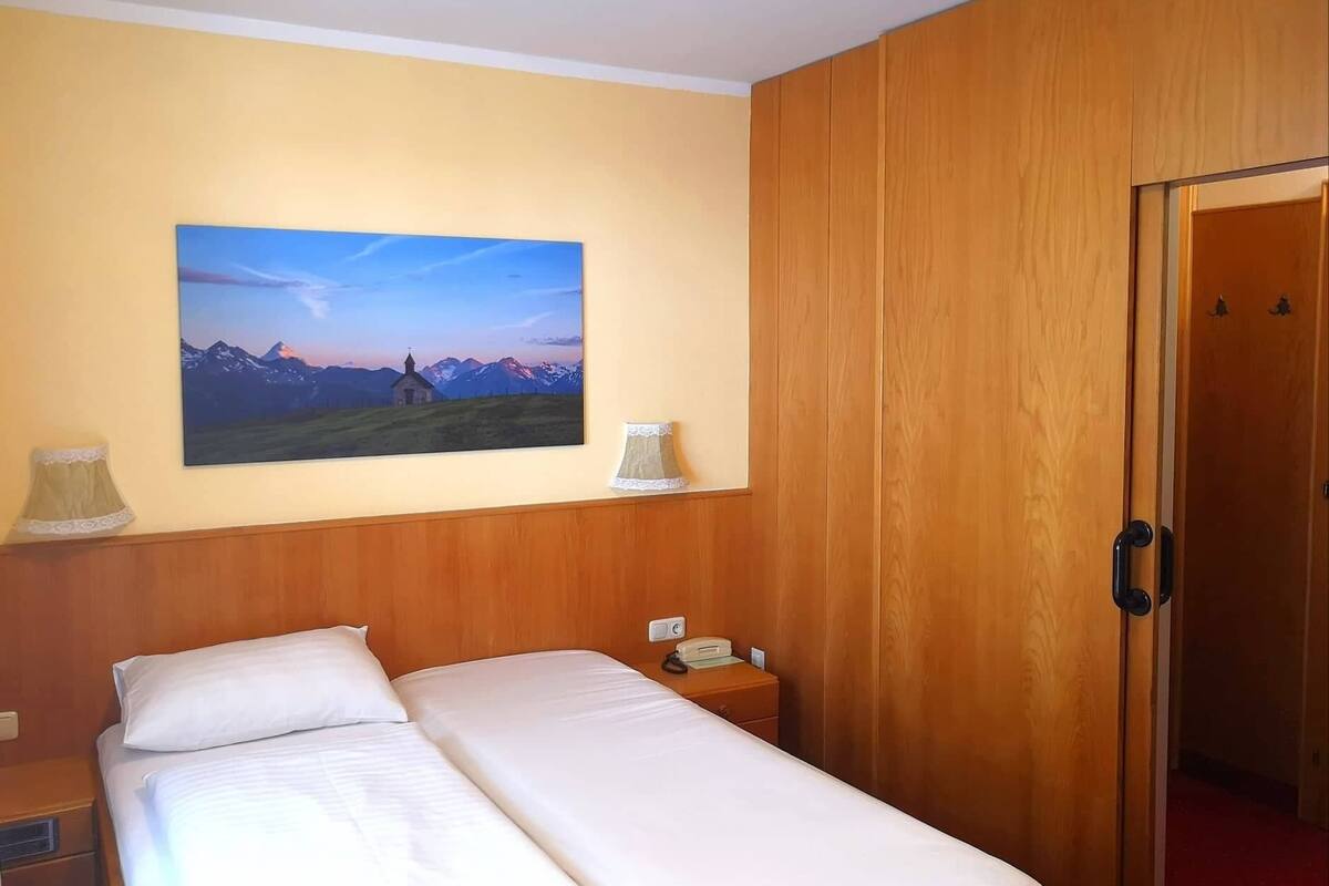A hotel room with a bed, a nightstand with a lamp, a painting of mountains, and a wooden door.