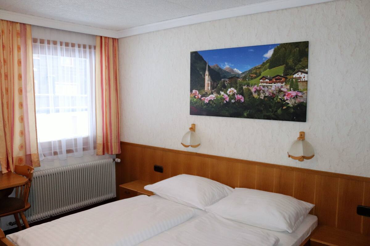 A hotel room with a bed, a window with curtains, a radiator, a table, and a wall-mounted picture of a landscape.