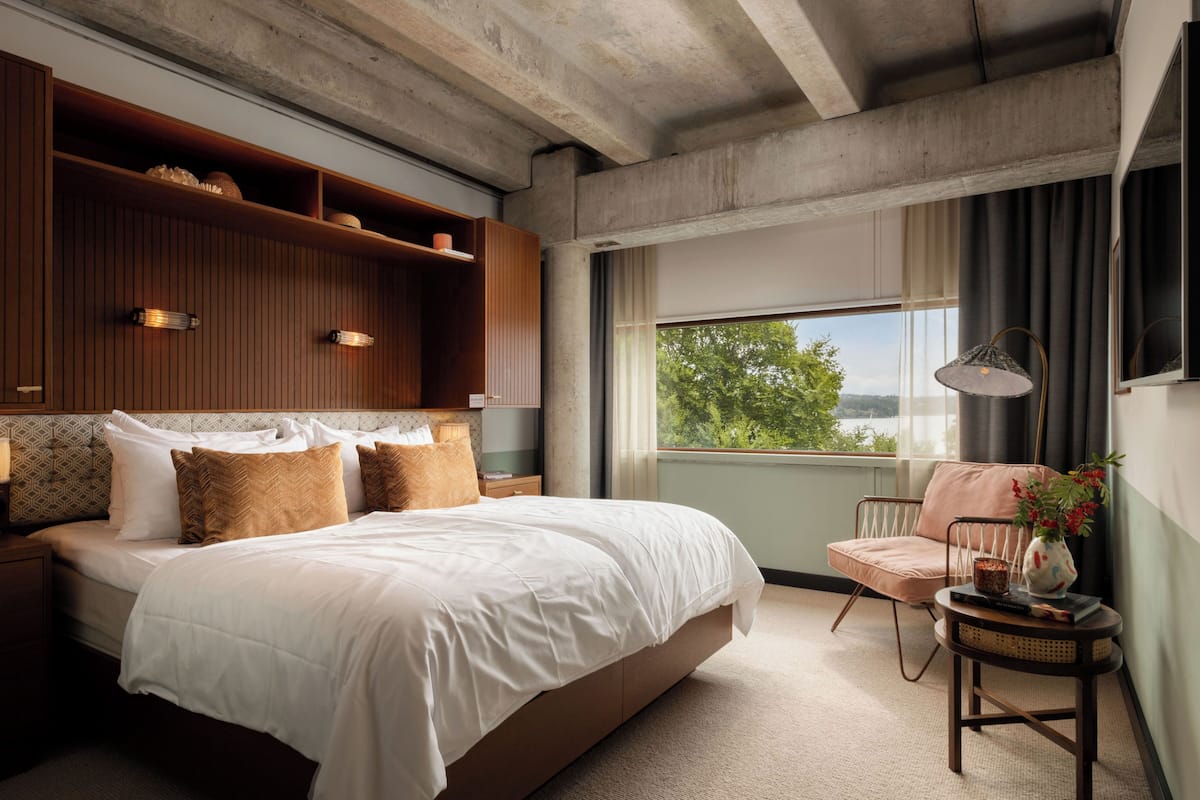 Une chambre moderne avec un grand lit, des étagères en bois, une vue sur la verdure et un coin salon confortable.