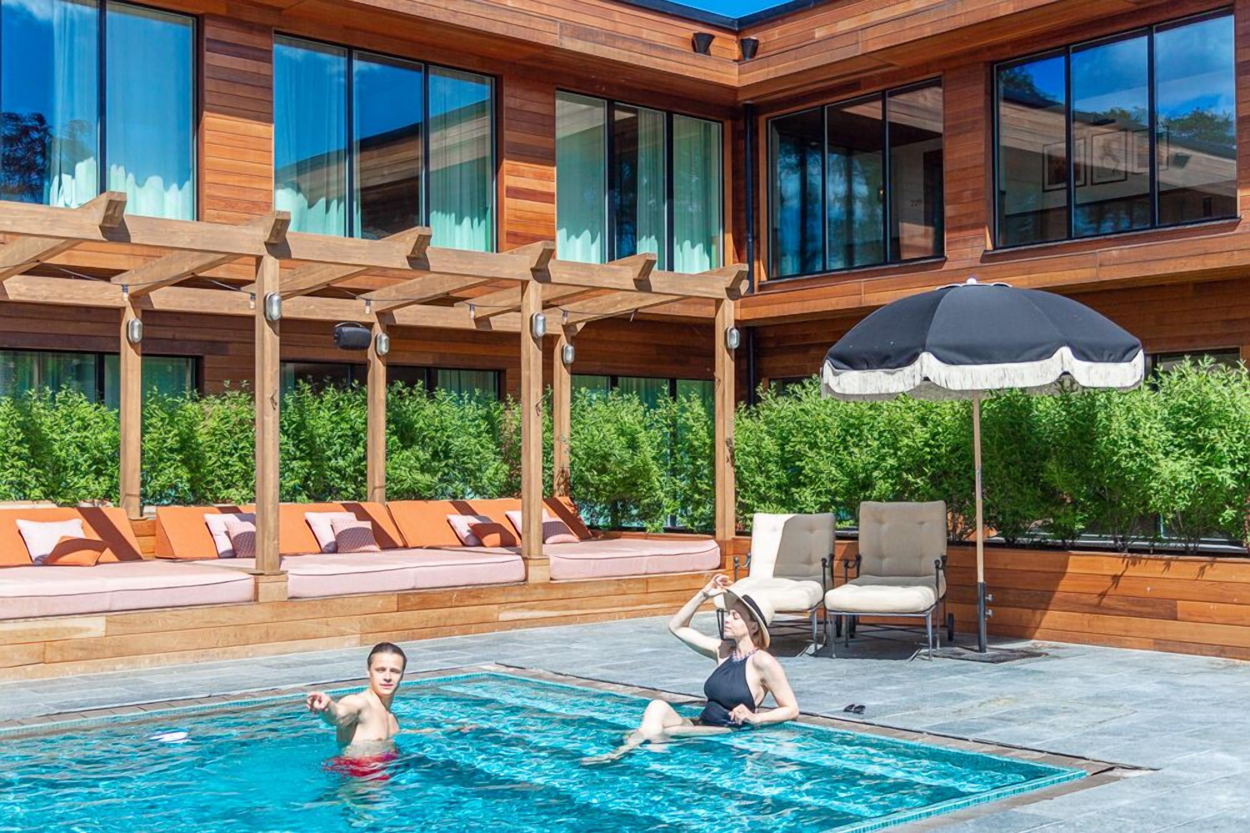 A modern outdoor pool area with wooden pergolas, lounge seating, and a large umbrella.