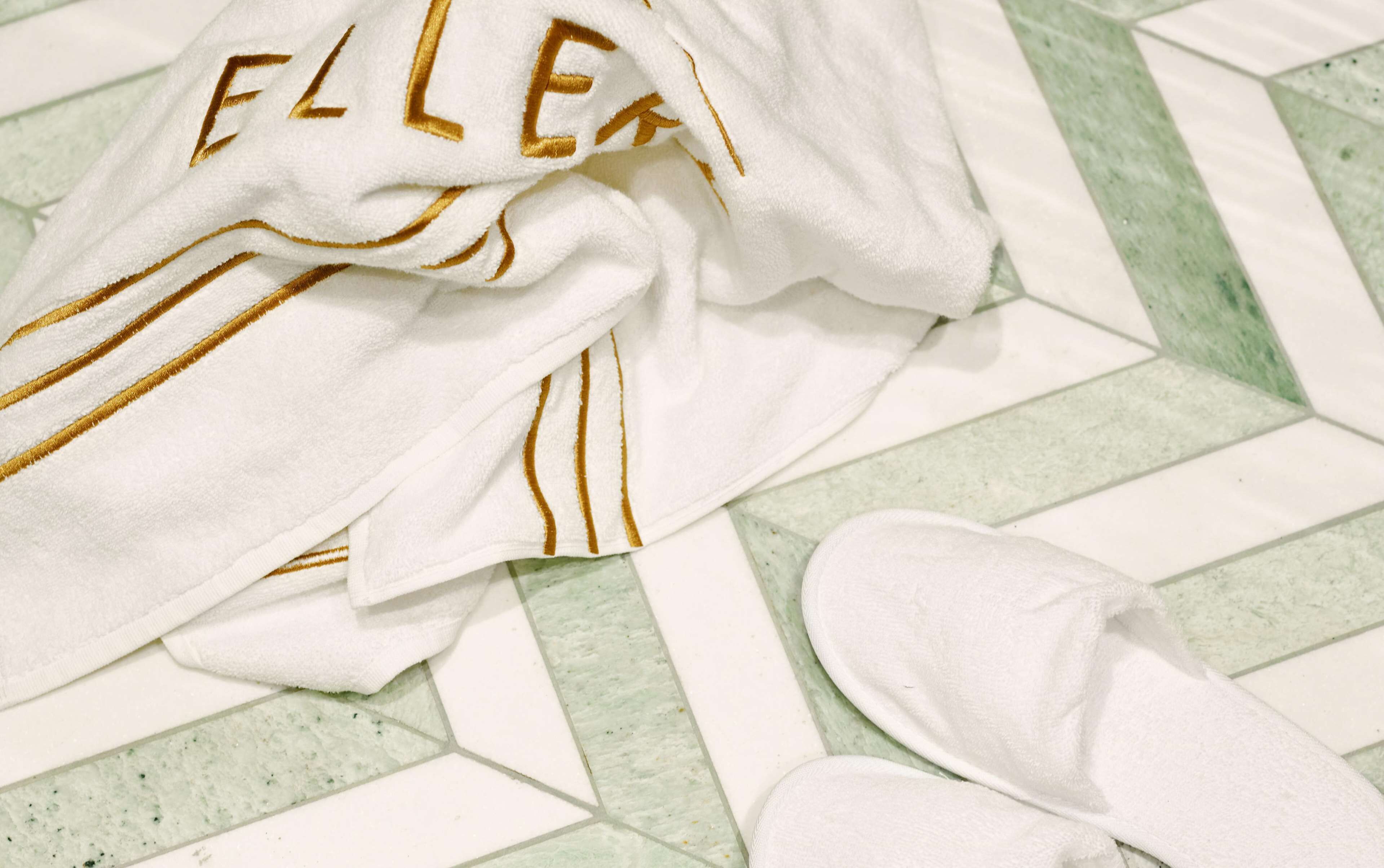 A white towel with gold lettering and a white slipper on a tiled floor.