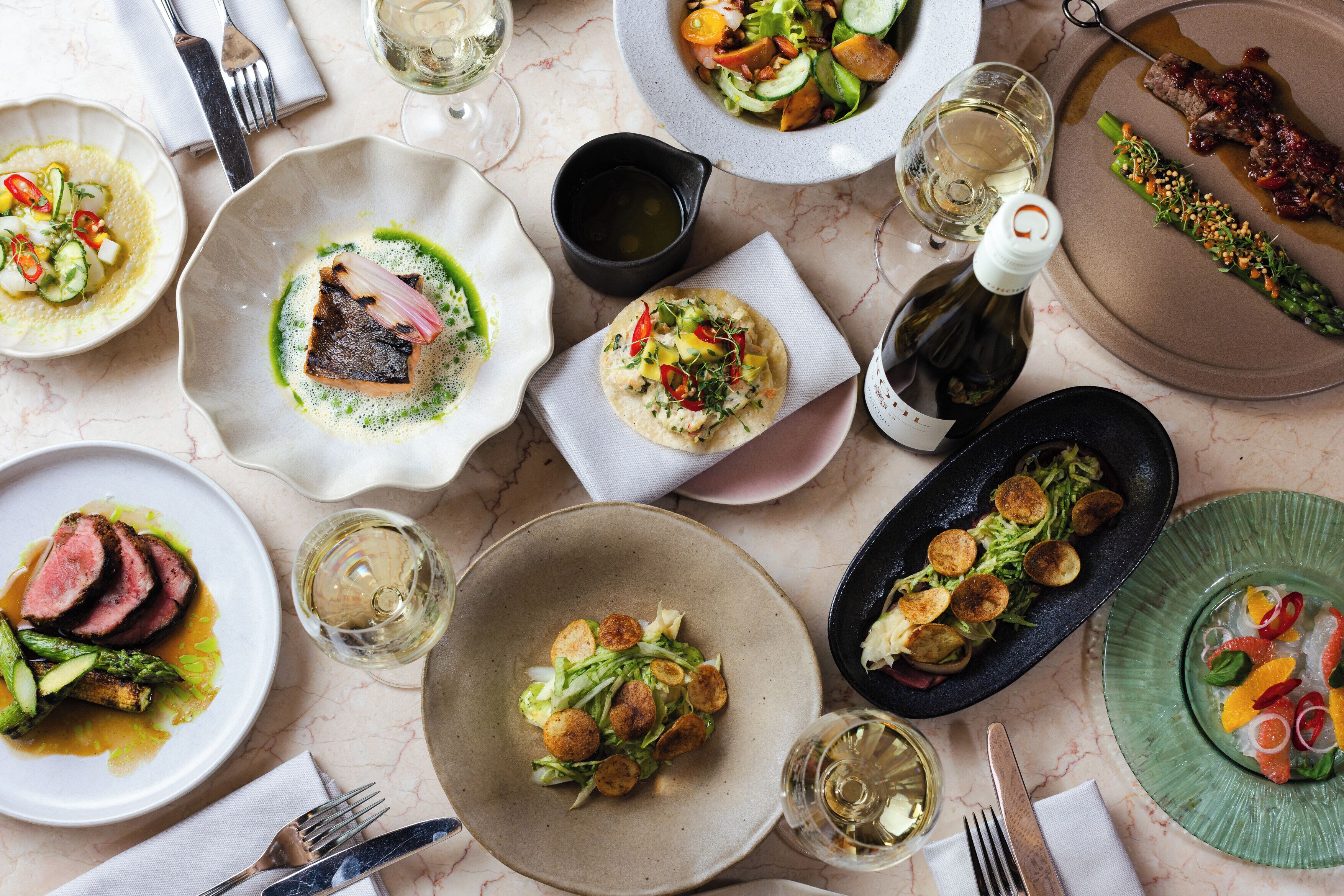 A variety of dishes on plates, a bottle of wine, and cutlery on a marble table.