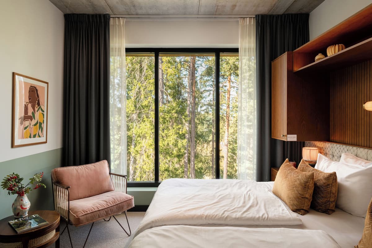 Une chambre avec une grande fenêtre, un lit recouvert de draps blancs, une chaise rose et une étagère en bois ornée d’objets décoratifs.