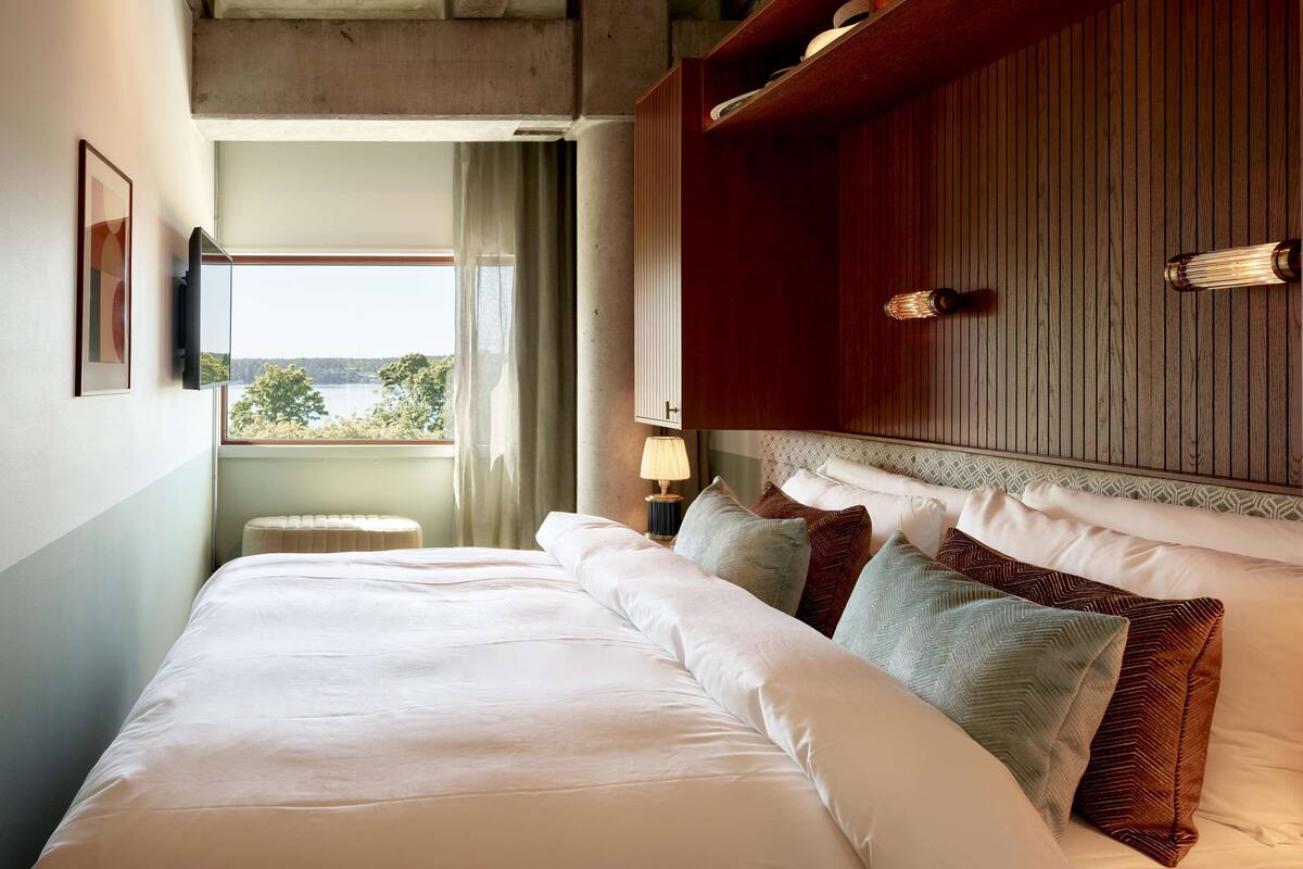 Une chambre d’hôtel avec un grand lit, une tête de lit en bois, une lampe de chevet et une vue sur les arbres par la fenêtre.