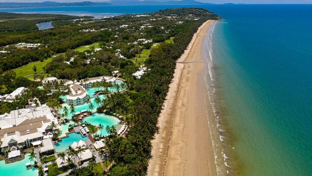 Eine Luftaufnahme eines Küstenresorts mit einem langen Sandstrand, einem großen Poolbereich und üppiger Vegetation.