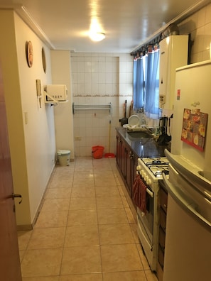 Una cocina con estufa a gas, horno y refrigerador. Se observa una ventana con cortinas azules, un lavamanos y un cubo de basura.