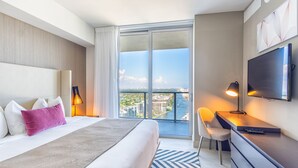 A hotel room with a large bed, a desk, a TV, and a view of the city through a large window.