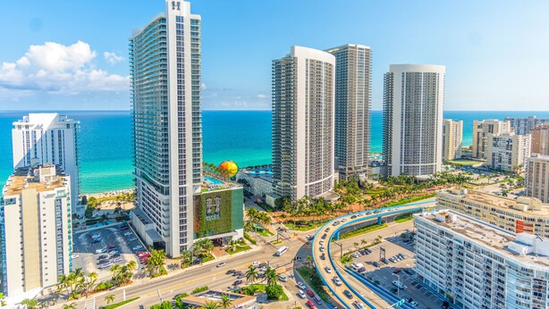 A high-rise hotel with a view of the ocean, featuring a curved road and palm trees.