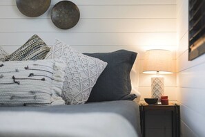 Una cama bien tendida con almohadas estampadas, una lámpara de noche y una mesita con un tarro y un cuenco.