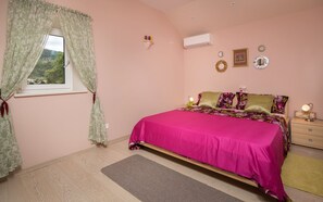 A bedroom with a bed, a window with curtains, an air conditioner, a bedside table with a lamp, and a small shelf with decorative items. - Villa Jasmine (Stari Grad)