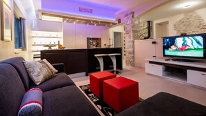 A modern living room with a sofa, red ottomans, a TV, and a kitchen area in the background. - Villa Jasmine (Stari Grad)
