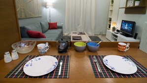 A dining area with a table set for two, a sofa, a TV, and a small coffee table with books and a lamp.