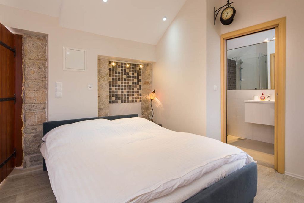 A bedroom with a bed, a stone wall, a clock, and a bathroom visible through an open door.
