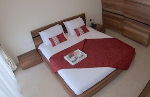 A neatly made bed with a red blanket and pillows, a tray with cups and a book, and a wooden dresser in the room.