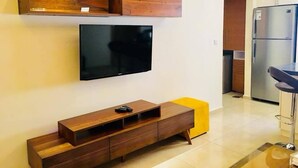 A modern living room with a flat-screen TV mounted on the wall, a wooden entertainment unit below, and a yellow ottoman. - Pearl Hotel & Residence (Freetown)