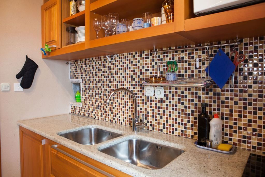 A kitchen with two stainless steel sinks, a tiled backsplash, wooden cabinets, and a shelf with glassware and kitchen items.