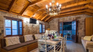 A cozy room with a stone wall, wooden ceiling beams, a dining table with chairs, a sofa, and a view through a window. - Studio Zavala (Jelsa)
