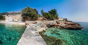 A coastal scene with a stone pathway leading to a rocky shore, clear turquoise water, and a building with a red roof. - Studio Zavala (Jelsa)