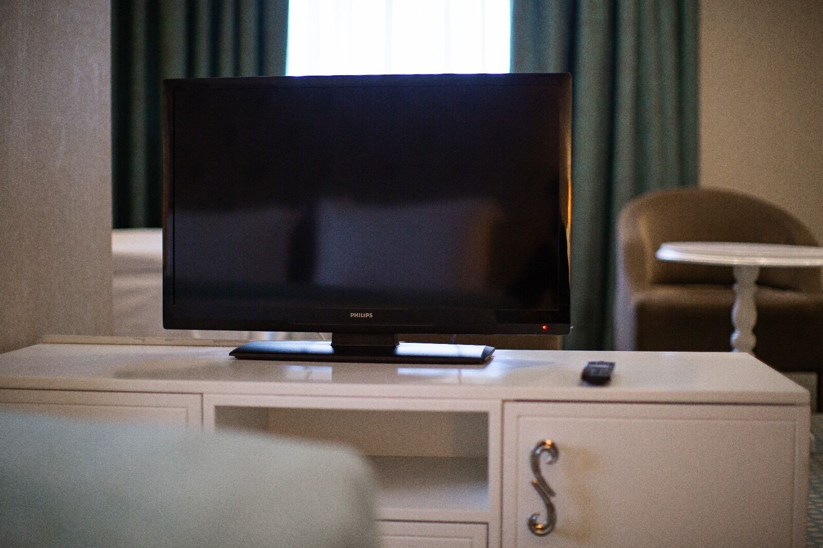 Quarto de hotel com televisão de ecrã plano sobre uma mesa consola branca, um comando e uma cadeira.