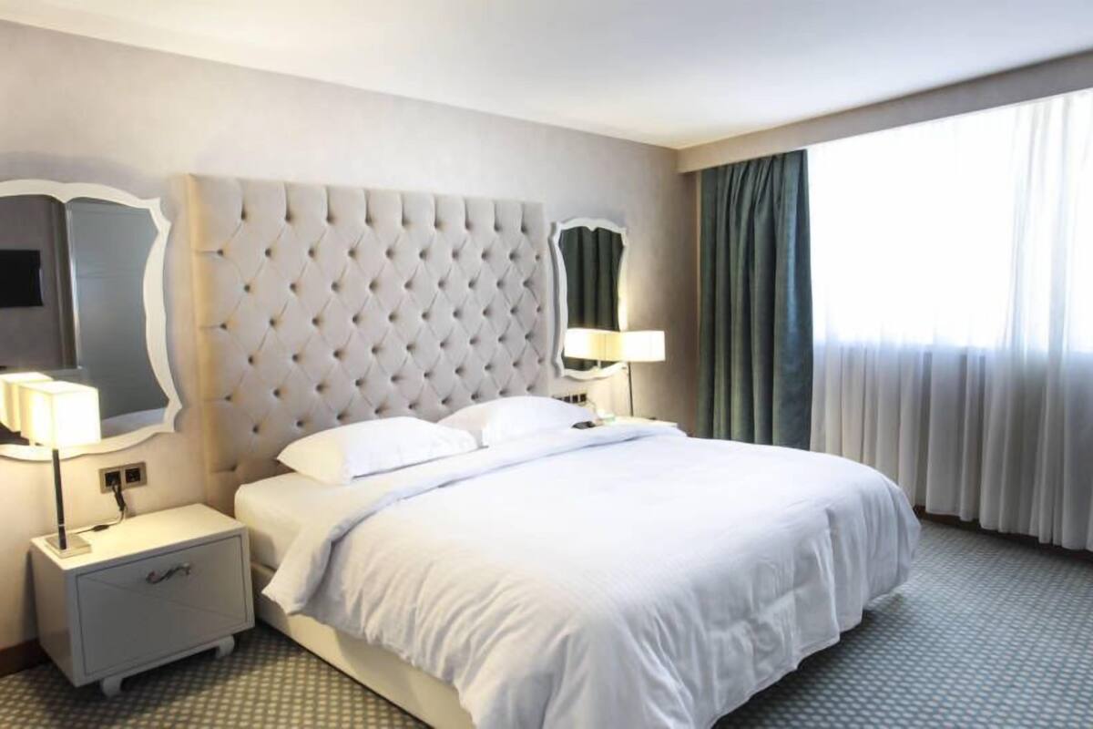 Ein Hotelzimmer mit einem geblümten Kopfstück, einem großen Bett, einem Nachttisch mit Lampe, einem Spiegel und einem Fenster mit Vorhängen.