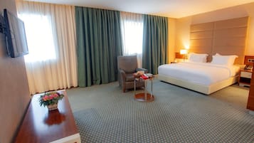 Une chambre d’hôtel avec un lit, une chaise, une petite table sur laquelle est posé un vase de fleurs, et un téléviseur fixé au mur.