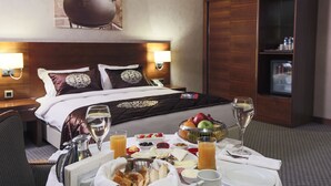 A hotel room with a neatly made bed, a breakfast table with fruit, juice, and a bottle of wine, and a minibar. - Nevali Hotel (Sanliurfa)