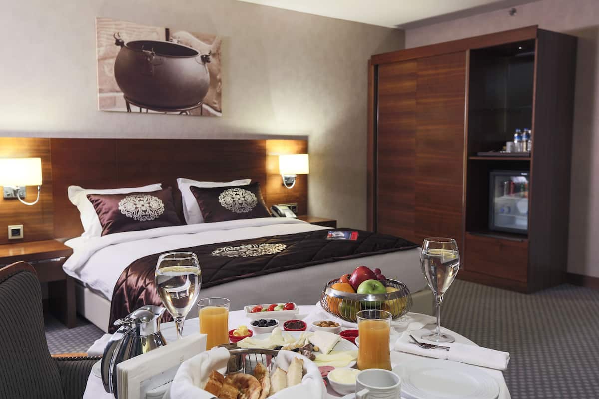 Ein Hotelzimmer mit einem ordentlich bezogenen Bett, einem Frühstückstisch mit Obst, Saft und einer Weinflasche sowie einer Minibar.