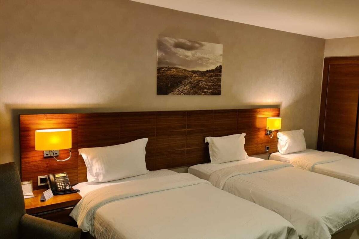 Ein Hotelzimmer mit zwei Betten, einem Holzkopfstück, Nachttischlampen, einer kleinen Kühlbox und einem Gemälde an der Wand.