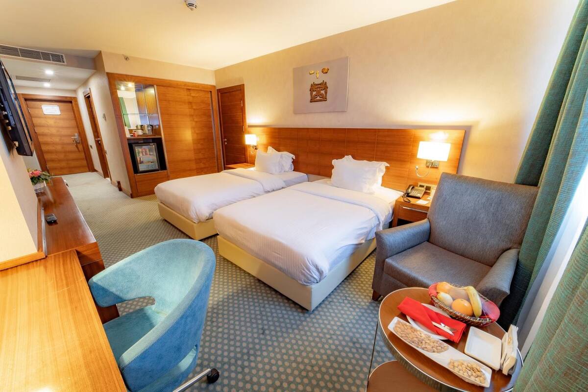 Ein Hotelzimmer mit zwei Betten, einem Schreibtisch, einem Stuhl, einem kleinen Tisch mit Snacks und einem Spiegel.