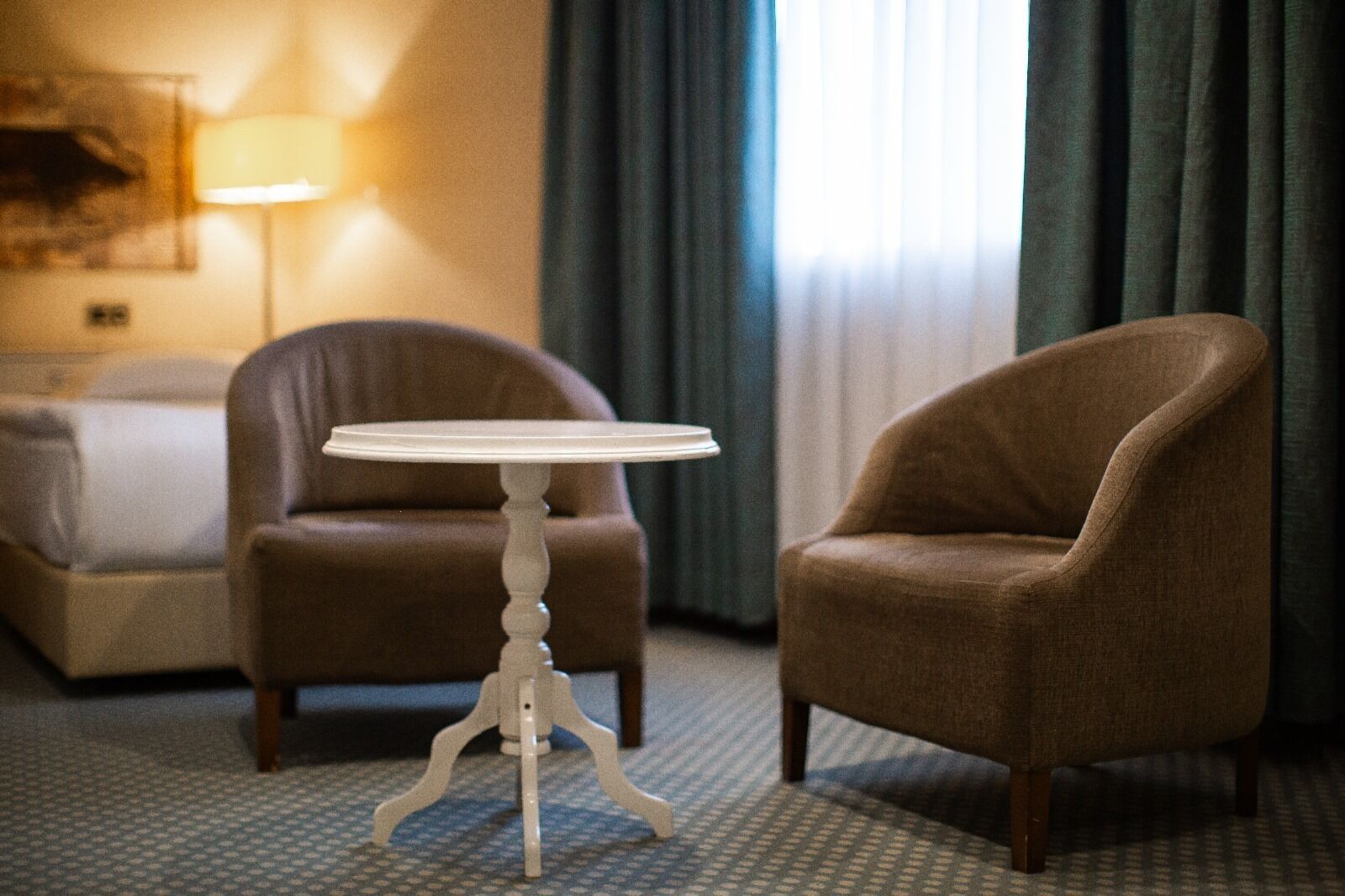 Una habitación de hotel con una cama, dos sillones, una mesita redonda, una lámpara y una ventana con cortinas.