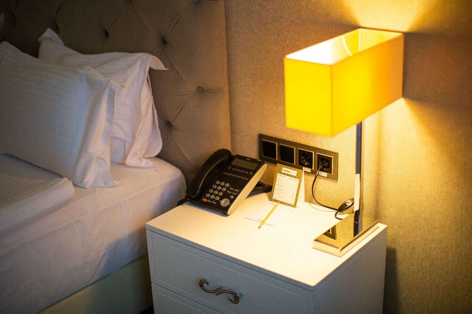 Cama bem arrumada com lençóis brancos, mesa de cabeceira com telefone, abajur e um pequeno cartão sobre a mesa.