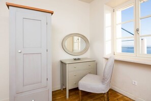 Um quarto com piso de madeira, um guarda-roupa branco, uma cômoda pequena e branca, um espelho redondo e uma janela com vista para o mar.