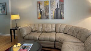 A living room with a beige sectional sofa, a small round coffee table with a magazine, a floor lamp, and two cityscape paintings on the wall.