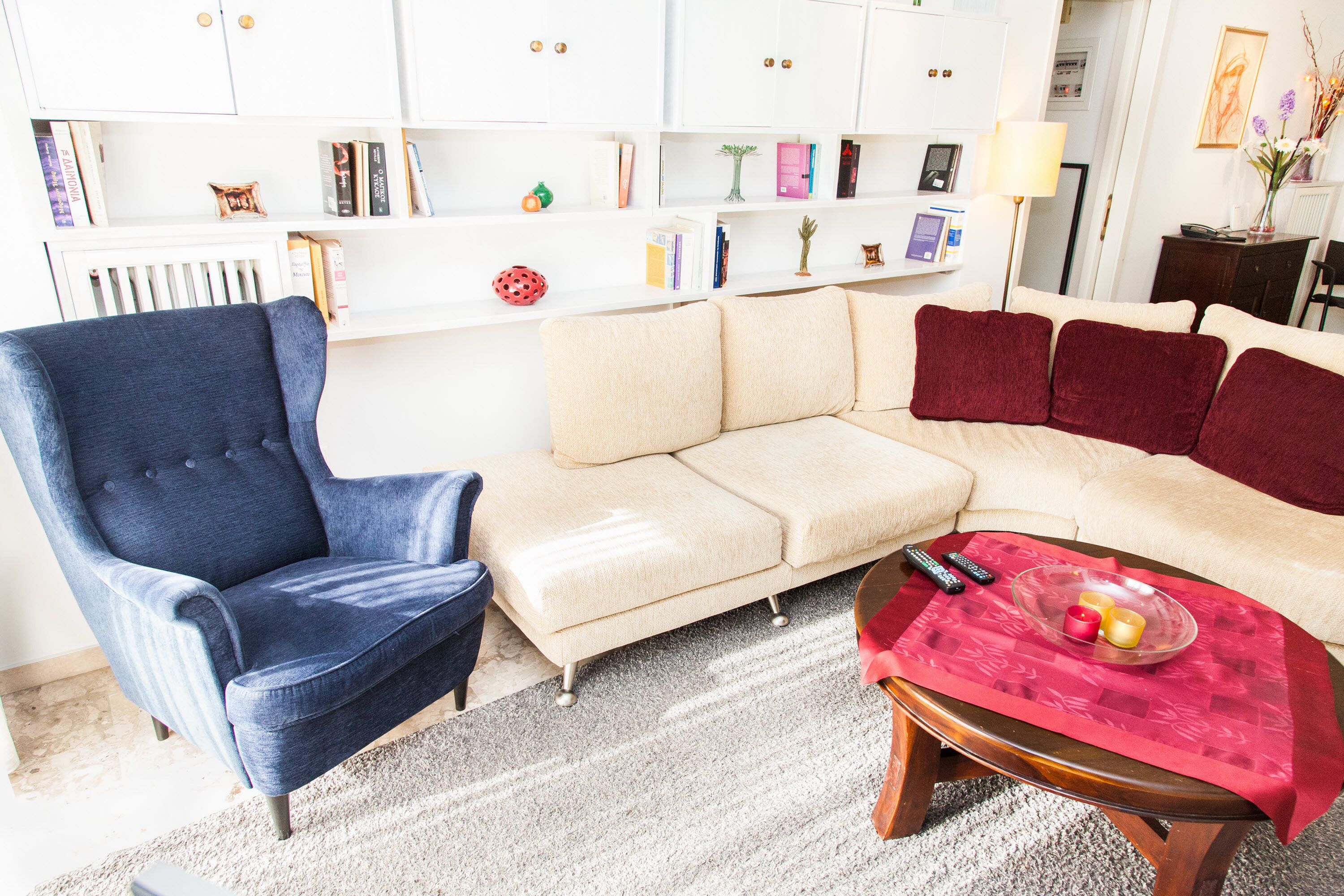 Una sala de estar con un sillón azul, un sofá color beige y una mesa roja con controles remotos.