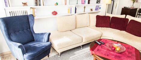 Una sala de estar con un sillón azul, un sofá color beige y una mesa roja con controles remotos.