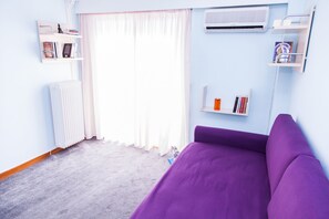 A room with a purple bed, white radiator, and a window with white curtains. There are bookshelves on the walls and a white air conditioning unit above the window. - Calm Oasis Apartment in Marousi (Marousi)