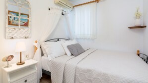 Ein ordentlich bezogenes Bett mit gesteppter Decke, ein Nachttisch mit Lampe, eine Wand-Klimaanlage und ein Fenster mit transparenten Vorhängen.