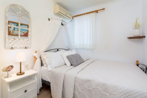 Een keurig opgemaakt bed met een gewatteerde hoes, een nachtkastje met een lamp, een wandairconditioner en een raam met dunne gordijnen.
