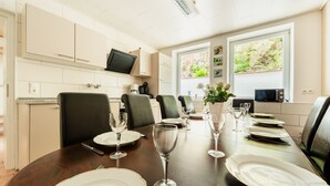 Un comedor con una larga mesa dispuesta para seis personas, con platos, cubiertos y copas de vino. Hay seis sillas, un televisor montado en la pared y una ventana con vistas a la vegetación.