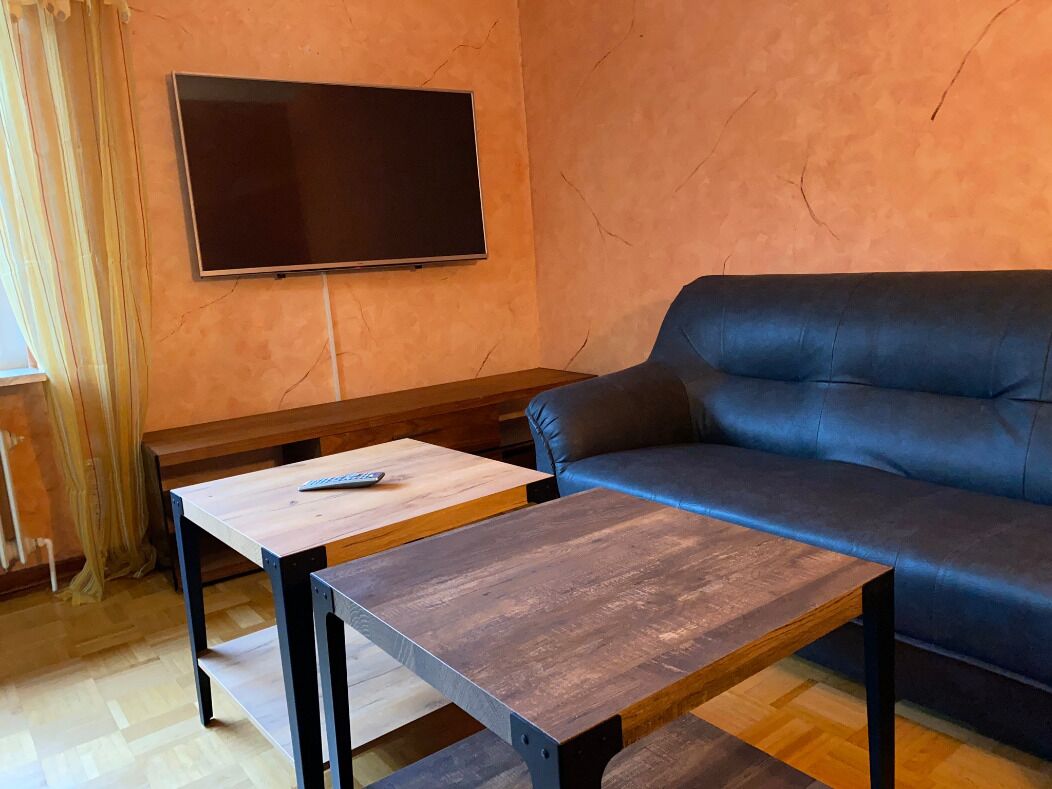 Una sala de estar con un sofá, dos mesas de madera, un televisor montado en la pared y una cortina a la izquierda.