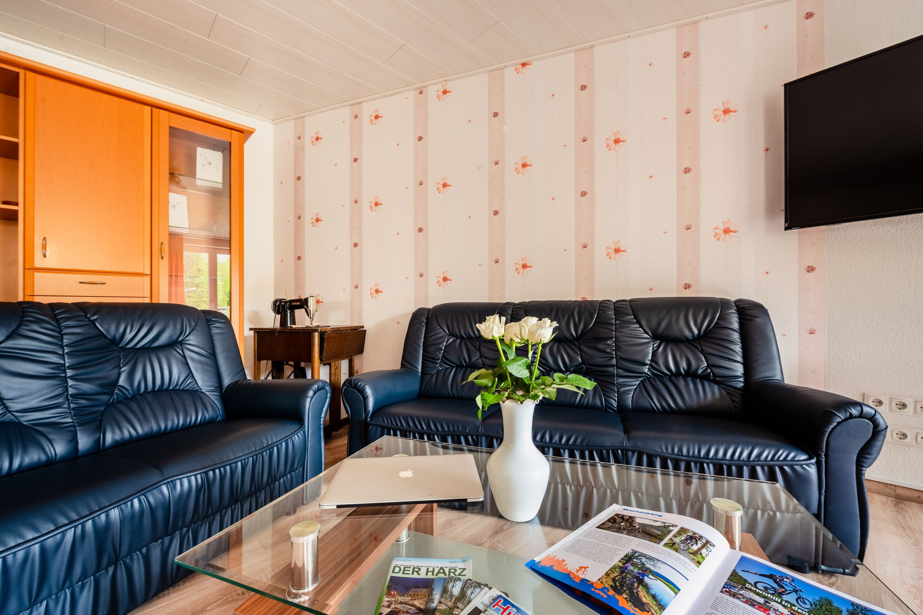 A modern living room with a glass coffee table, two black leather sofas, a white vase with flowers, and a laptop.