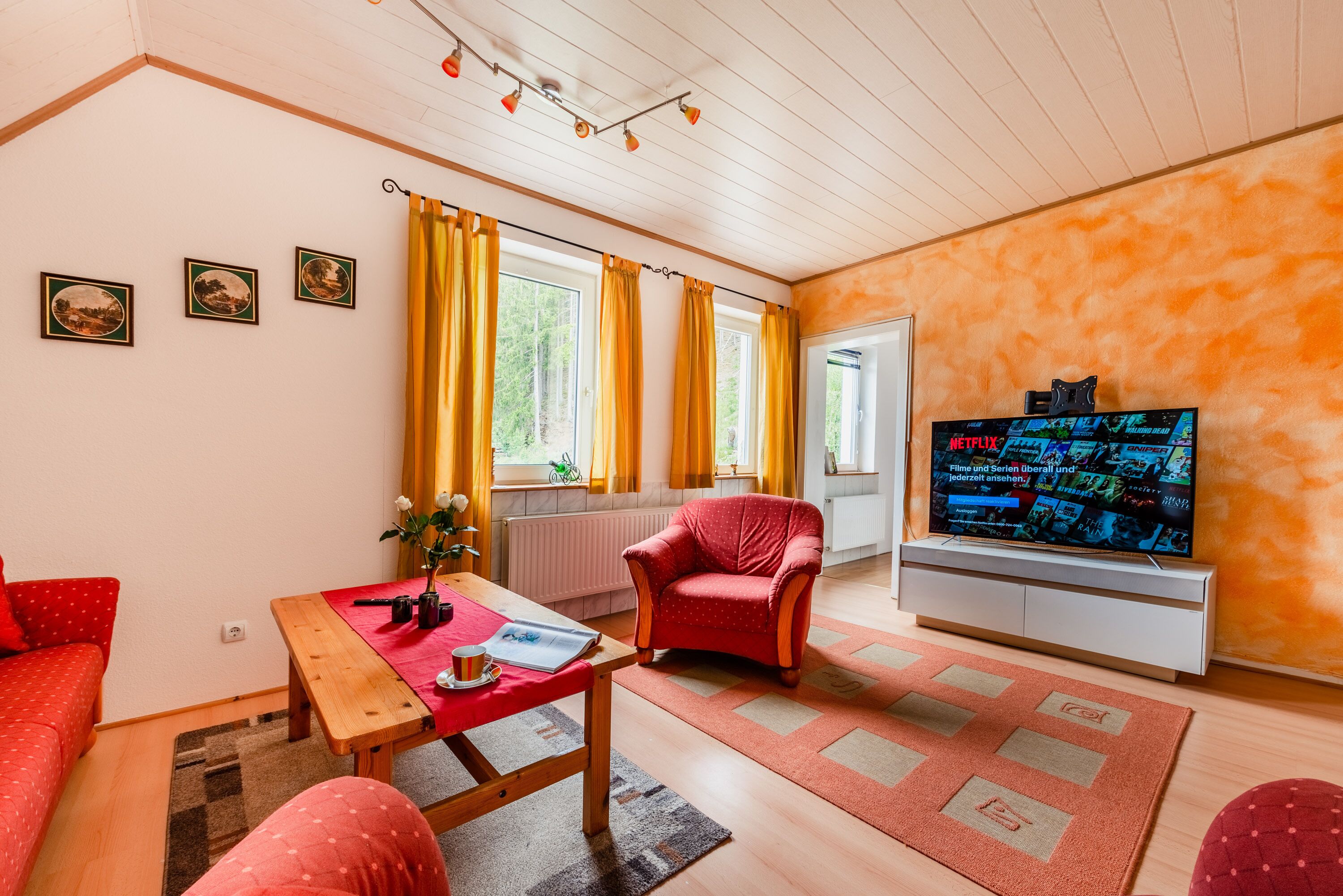 Una sala de estar acogedora con techo de madera, un sillón rojo, una mesa de madera y un televisor sobre un soporte blanco.