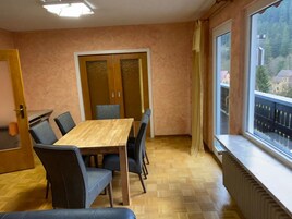 A room with a wooden dining table and chairs, a large window with a view of a building and trees, and a radiator on the wall.