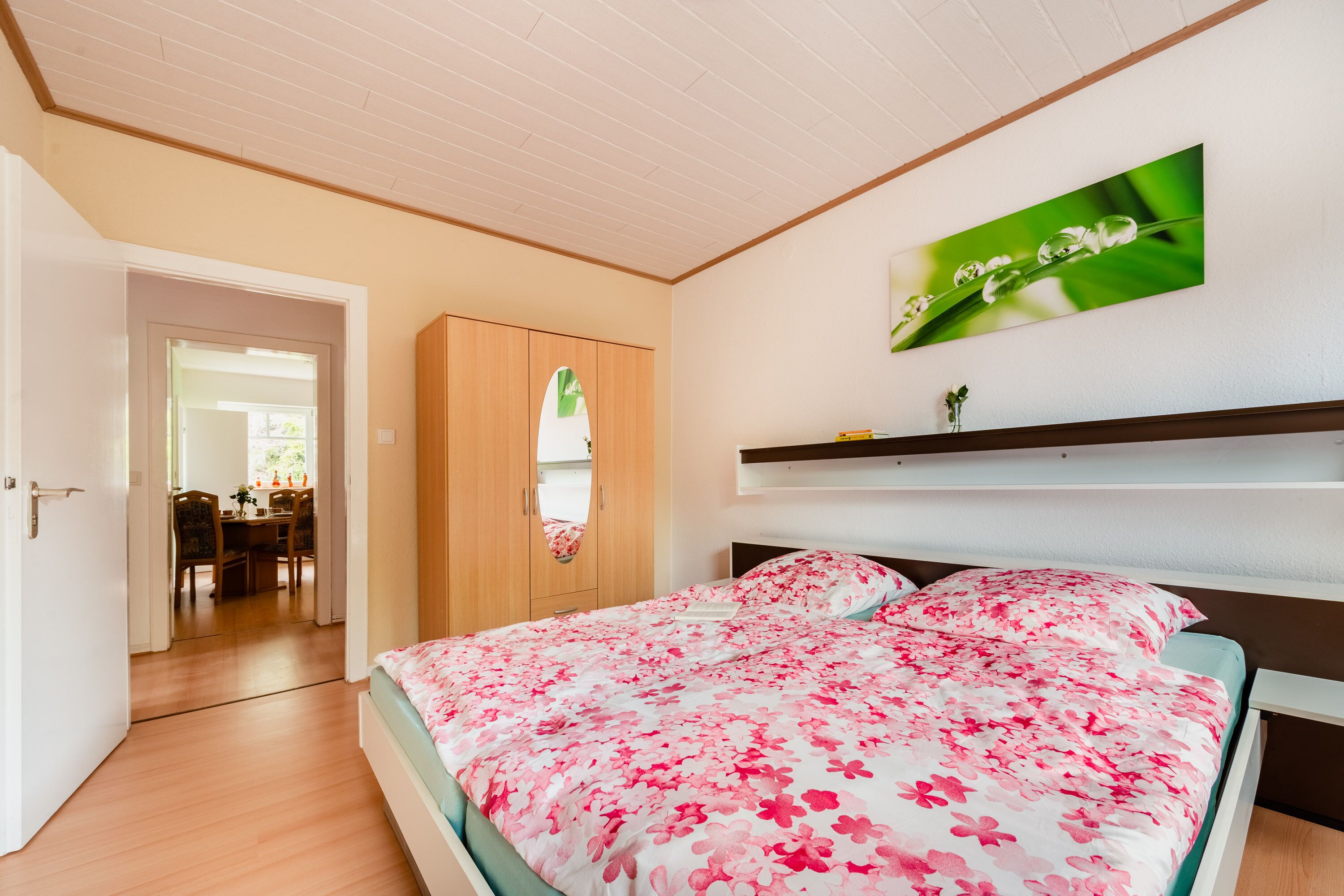 Un dormitorio con una cama, un armario de madera, un cuadro de pared verde y una zona de comedor visible a través de una puerta abierta.