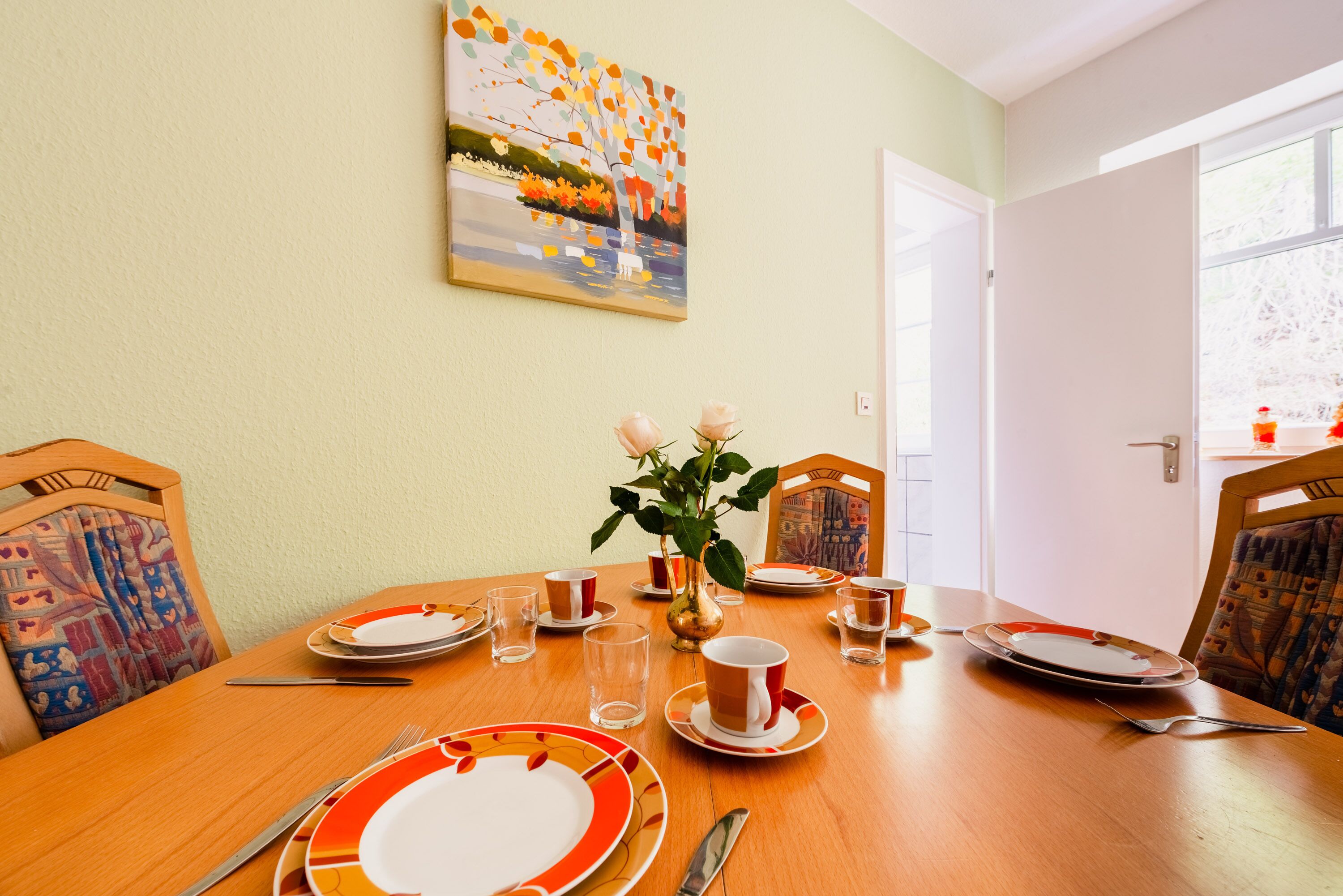Una mesa de comedor dispuesta con platos, tazas y cubiertos, un florero con flores y un cuadro en la pared.