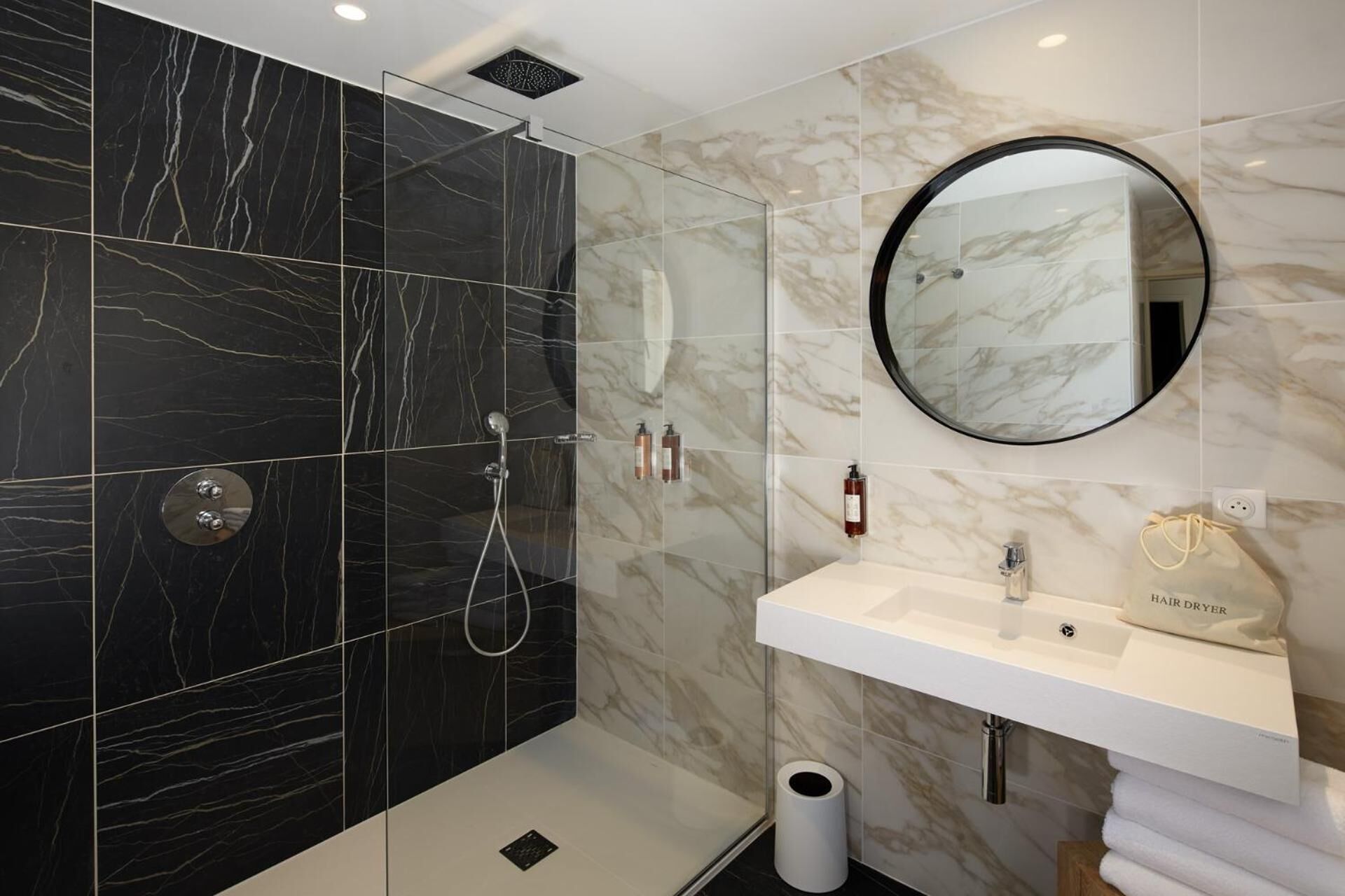A modern bathroom with a walk-in shower, a round mirror, a white sink, and a towel rack.